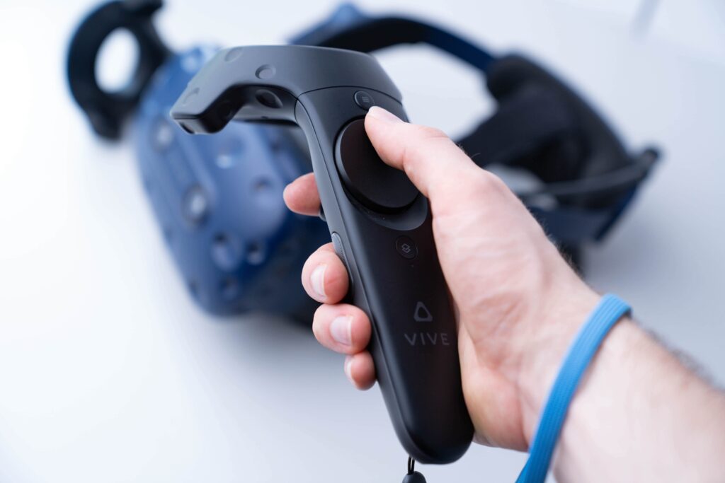 Ręka trzymająca kontroler VR marki Vive z goglami VR w tle